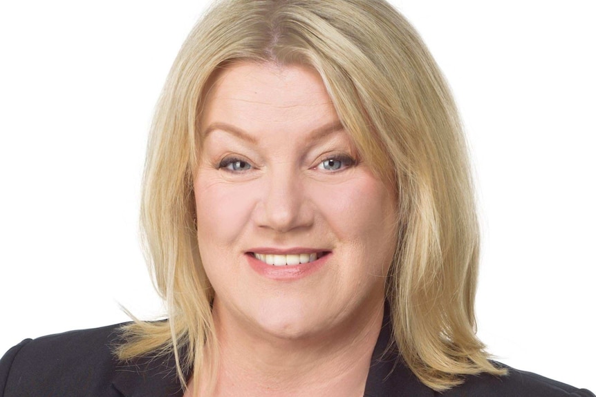 A woman with blonde hair wearing a dark jacket smiles at the camera in a posed portrait.