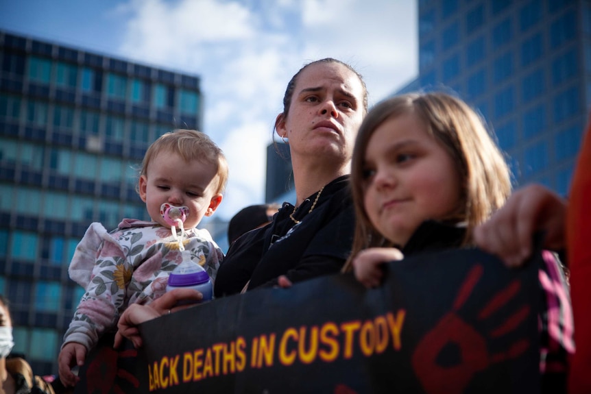 A woman holds a baby. She and a young girl also hold a sign reading 'black deaths in custody'