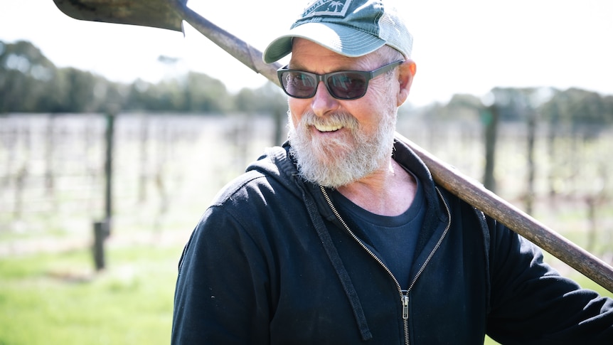 A man with a shovel over his shoulder in front of some vines