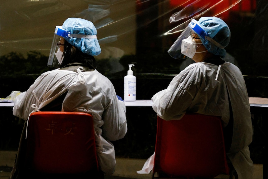 개인 보호 장비를 착용한 두 명의 의료 종사자가 보호된 방패 뒤에 나란히 앉아 있습니다.