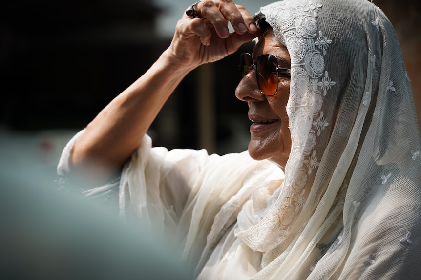 A woman wearing a white lace head scarf and sunglasses smiles slightly as she is photographed in profile, hand to her forehead
