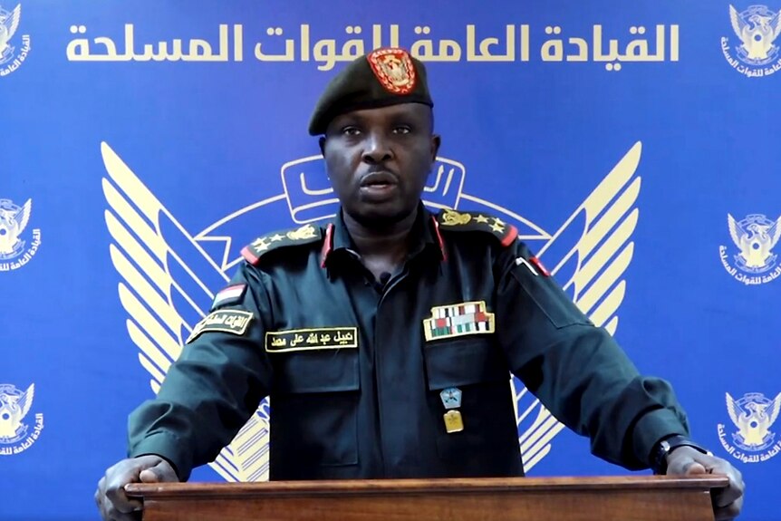 A middle-aged African man in a dark green miliatry uniform stands behind a lectern in front of a blue backdrop.
