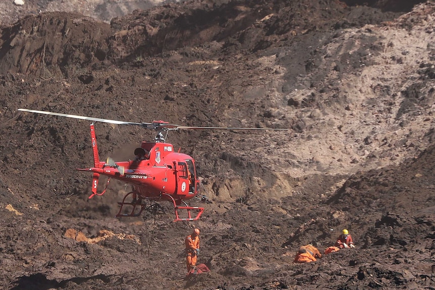 A helicopter hovers over rescue workers searching through muddy sludge