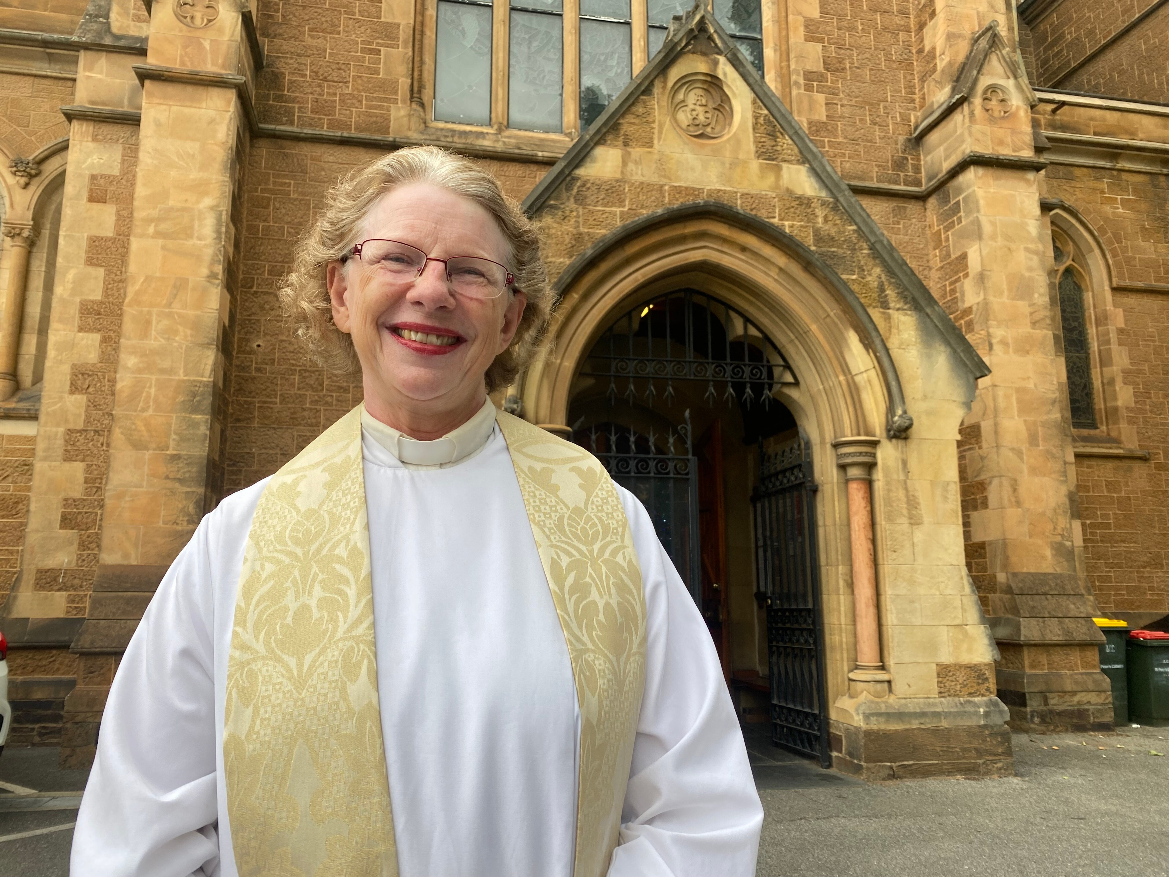 A woman with short blonde hair dressed in a white and gold robe smiles while standing in front of a church