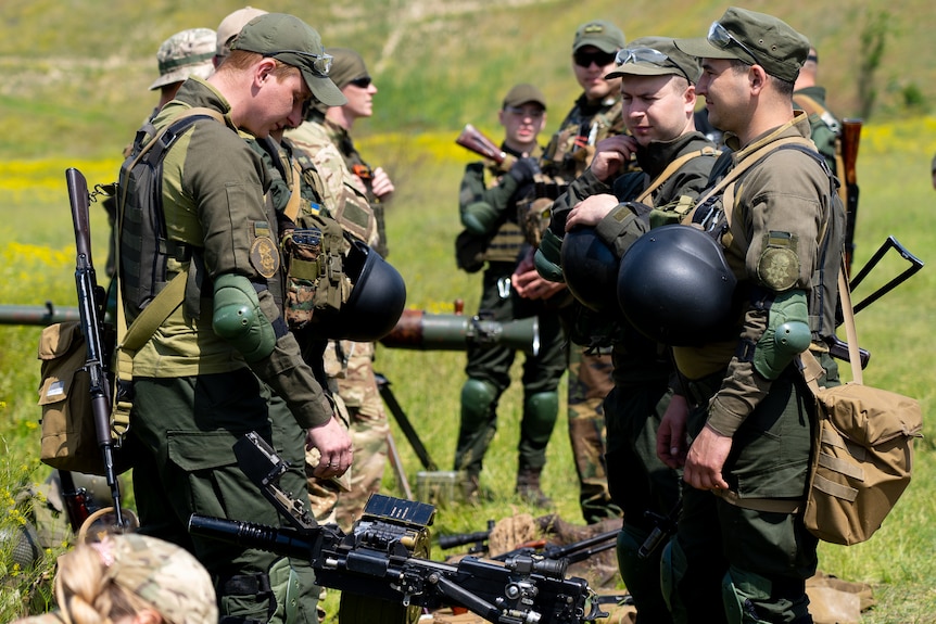 Men in camo gear and helmets stand around chatting while examining rifles 