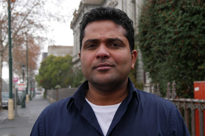 A man with dark hair and a blue collared shirt photographed on a street.