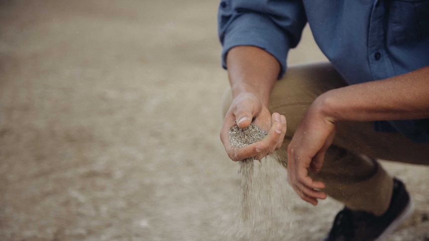 A man in farming gear kneels down and lets earth run through his fingers.