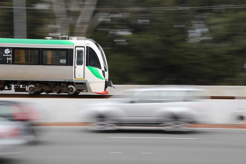 A Transperth train travels from left to right with blurry cars heading in the opposite direction in the foreground.
