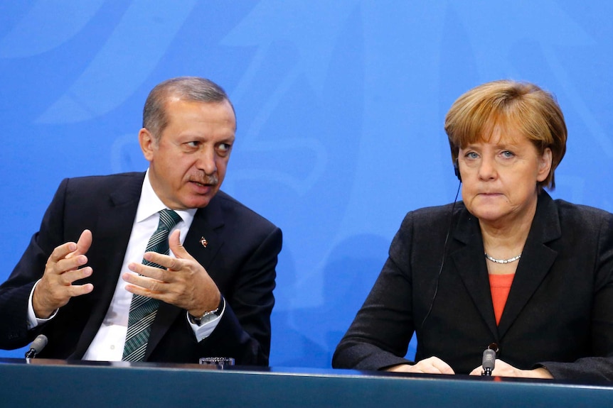 Tayyip Erdogan leans across to speak to a grim faced Angela Merkel.