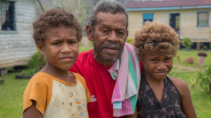 A man stands with his two grandchildren in front of damaged homes