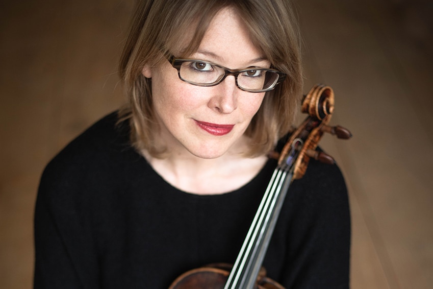 A woman sitting dressed in black cradles a violin in her lap and, with a neutral expression, looks up towards the camera.