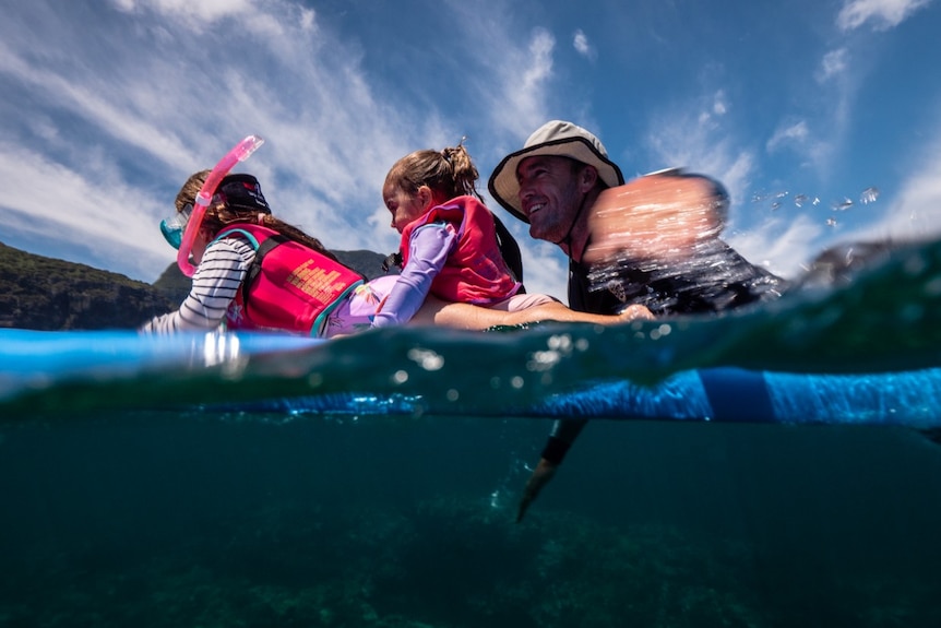 A man paddles on a surfboard with as his two young daughters lay in front of him.