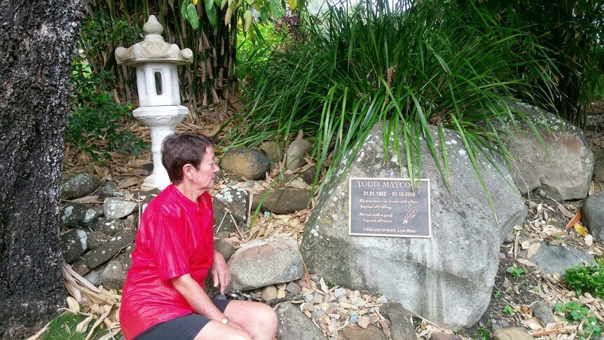 A woman sits in a garden looking at a plaque commemorating her son Todd.