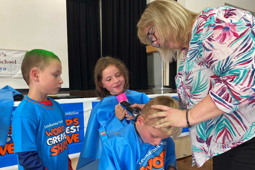 Two children in world's greatest shave clothes help a woman shave hair off a third child's head while they sit on a stool.
