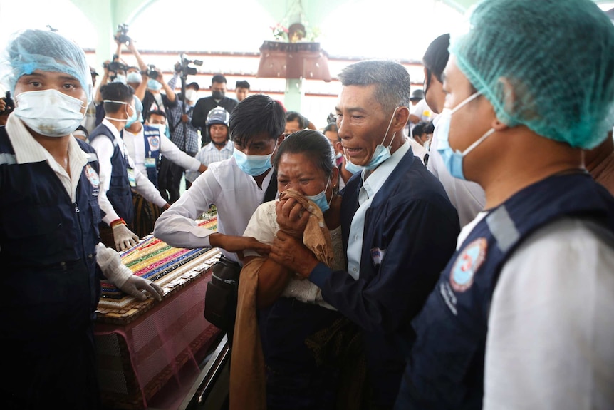 A crying woman is supported by two men as a coffin is carried through a dense crowd of people wearing face masks.