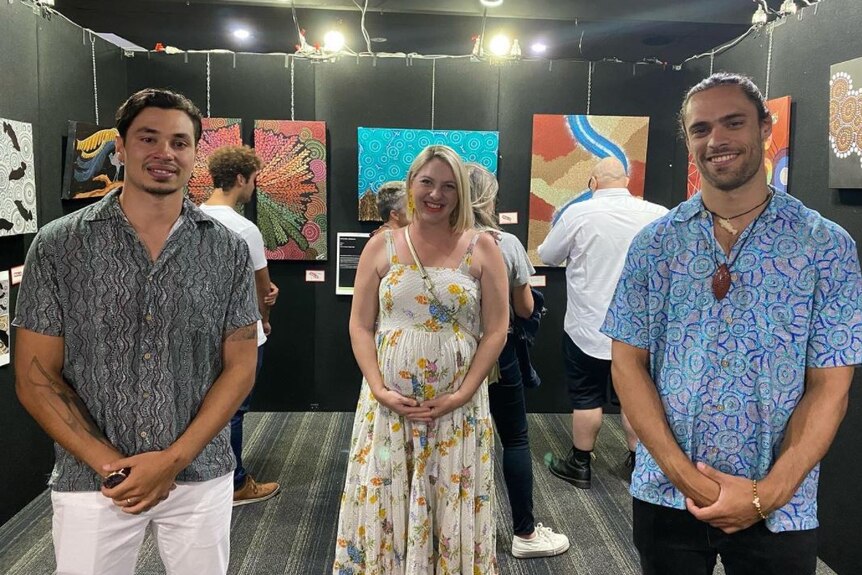 A pregnant smiling lady in a white dress stands in the middle of two men wearing in a gallery of Aboriginal art