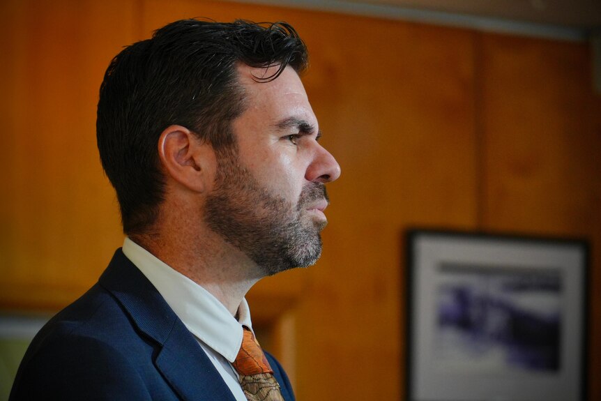 The side profile of a man in a blue suit with short dark hair and a small beard. He looks serious.
