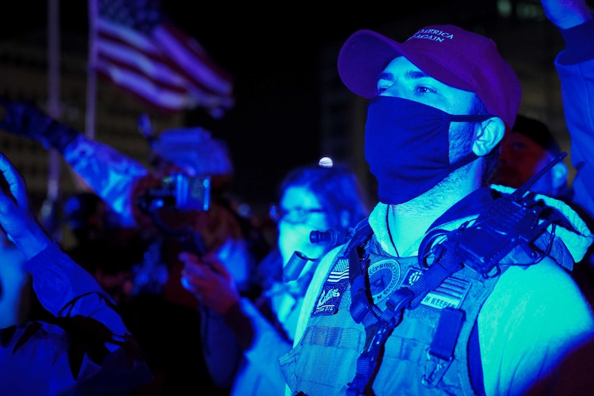 A man in a Trump cap and mask wearing a fatigue vest with a walkie talkie