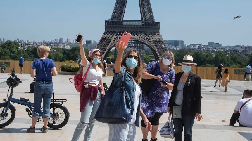 A group of people wearing facemasks hold their phones high to take a selfie on Trocadero square in front of the Eiffel Tower