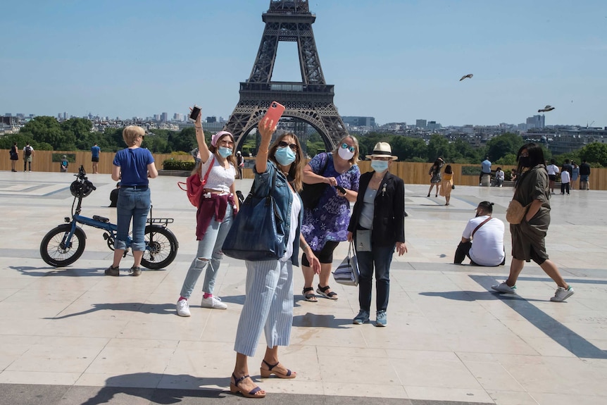 A group of people wearing facemasks hold their phones high to take a selfie on Trocadero square in front of the Eiffel Tower