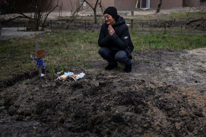 Tanya kneels by her husband's grave. On top is a small cross and some small momentos