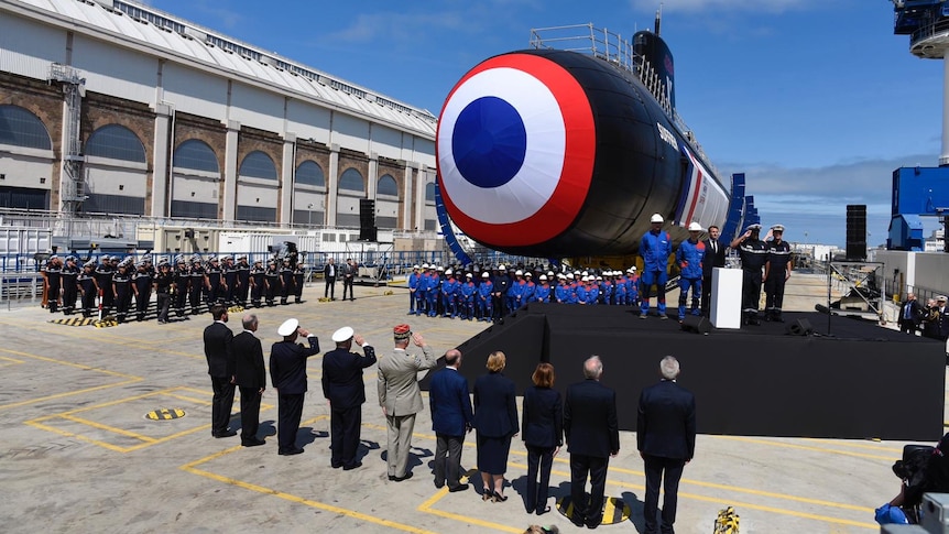 Navy officers and politicians salute a submarine with a target symbol painted on its front at a shipyard.