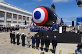 Navy officers and politicians salute a submarine with a target symbol painted on its front at a shipyard.