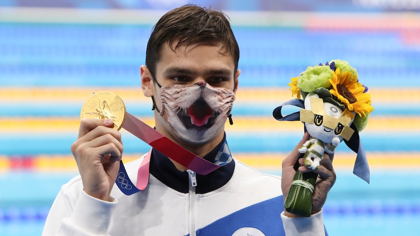 An Olympic champion swimmer stands at poolside holding his gold medal and a mascot in the other, wearing a tiger mask. 