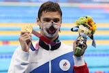 An Olympic champion swimmer stands at poolside holding his gold medal and a mascot in the other, wearing a tiger mask. 