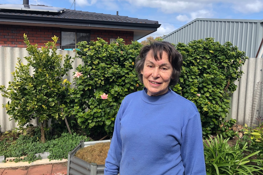 A woman in a blue sweater standing in her backyard in front of trees and a fence.