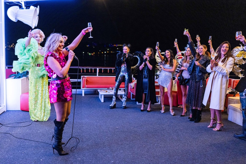 Kate Adams wears a pink sparkly dress and holds up a glass for a toast as she addresses a crowd on a boat.