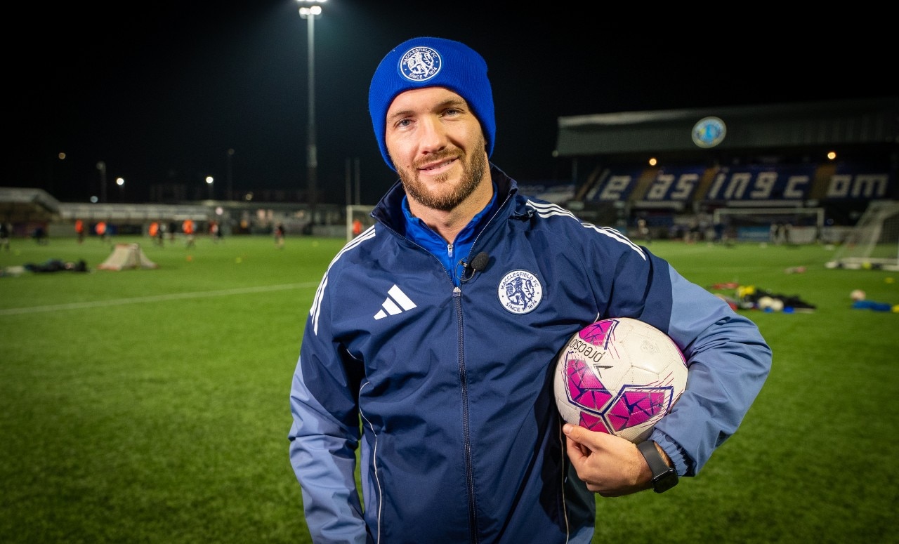 Um homem em traje esportivo olha para a câmera e sorri. Ele está em um estádio à noite.