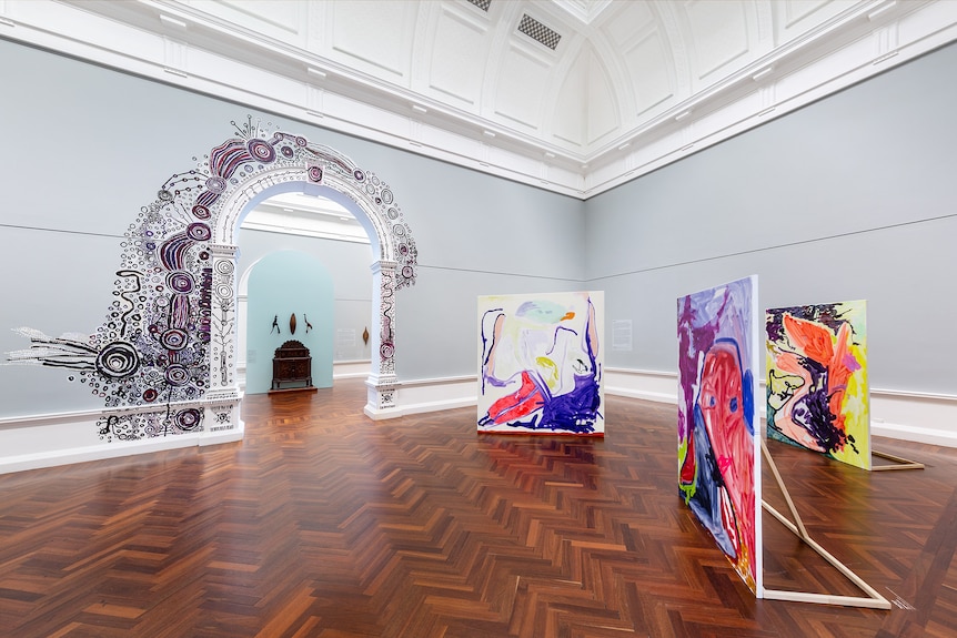 Gallery with wooden flooring and grey walls featuring three large colourful abstract paintings and site-work painted on an arch