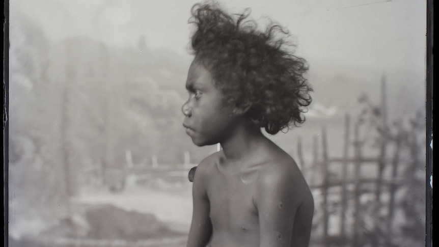 An Aboriginal child sits on a stool in a studio waiting to have his portrait photographed