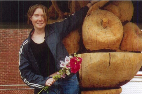 An aged photo of a young woman leaning against a wooden sculpture.