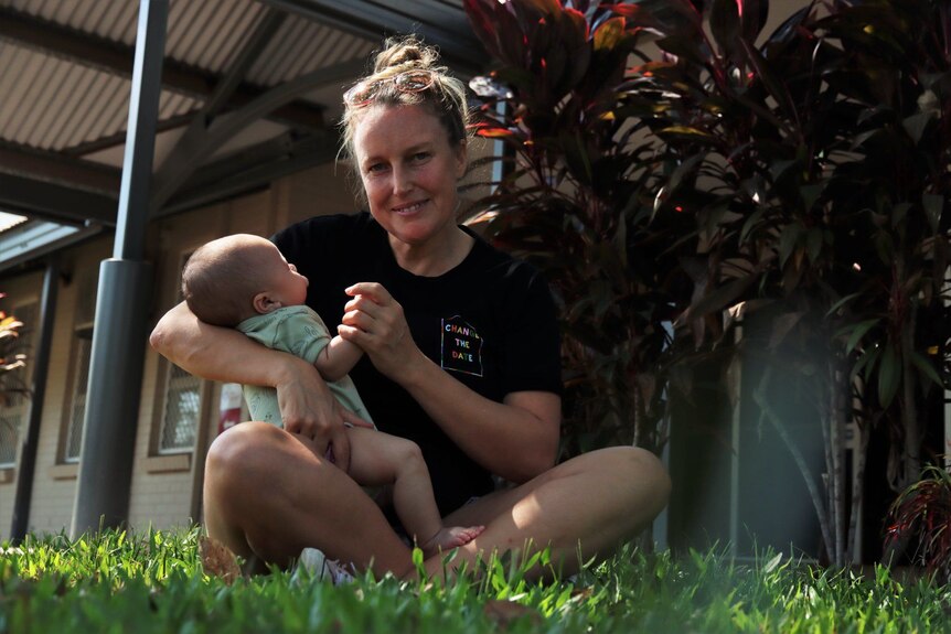 A mother with blonde hair in a bun wearing a black T-shirt is sitting on grass with a baby.