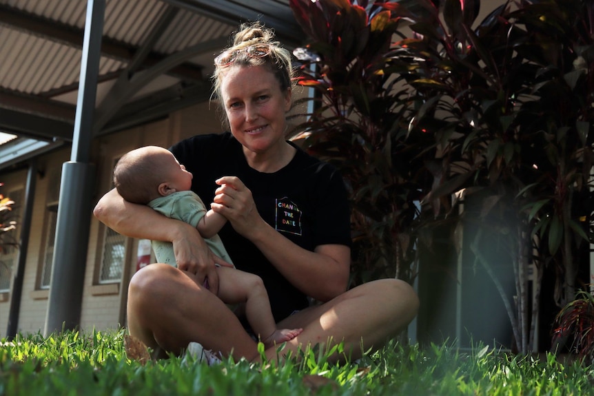 A mother with blonde hair in a bun wearing a black T-shirt is sitting on grass with a baby.