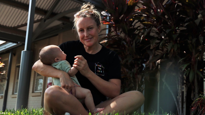 A mother with blonde hair in a bun wearing a black T-shirt is sitting on grass with a baby.