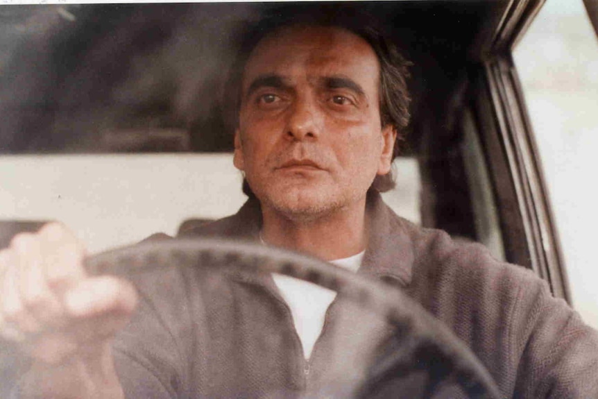 An older Iranian man in a car, he grips the steering wheel