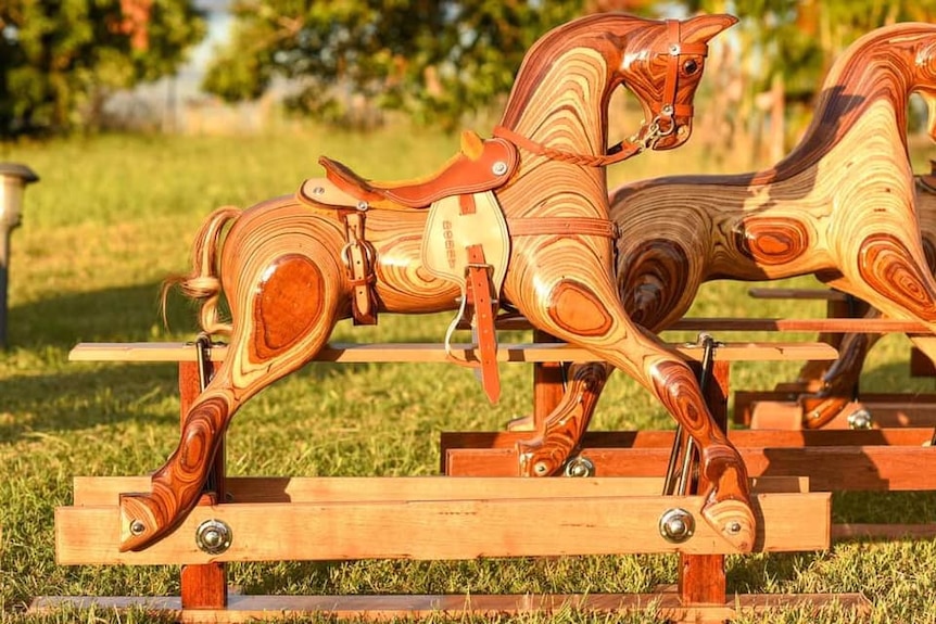 Two timber rocking horses with leather saddle out on the grass with greenery in the background.