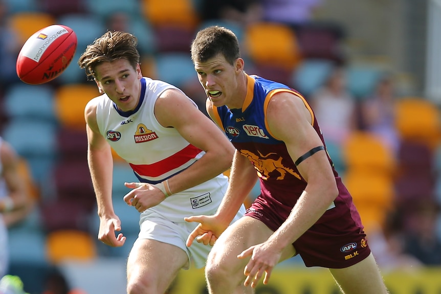 A Brisbane Lions AFL player eyes the ball as he runs after it, while a Western Bulldogs player looks across at him.