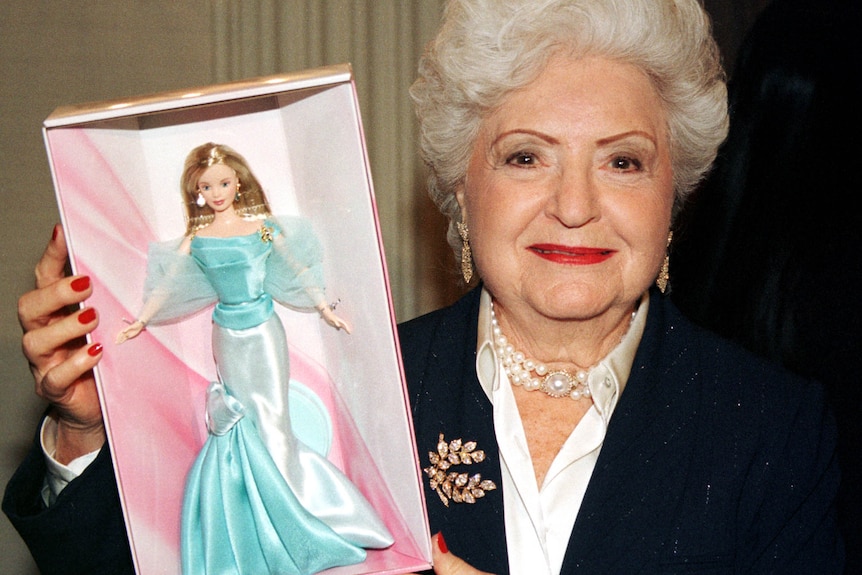 A woman with white hair and wearing a pearl necklance poses with a Barbie doll wearing a blue dress.