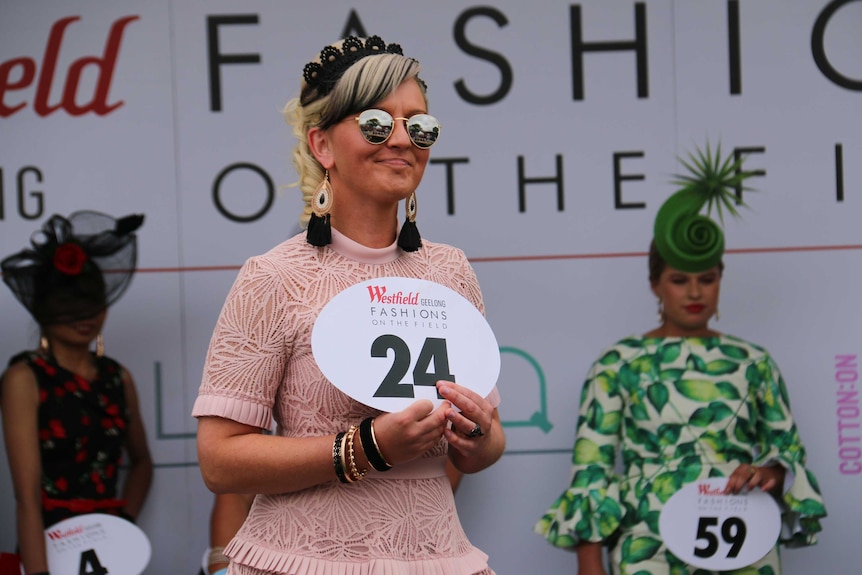 A woman on an outdoor stage, dressed in racing fashion, holds a big number and smiles.