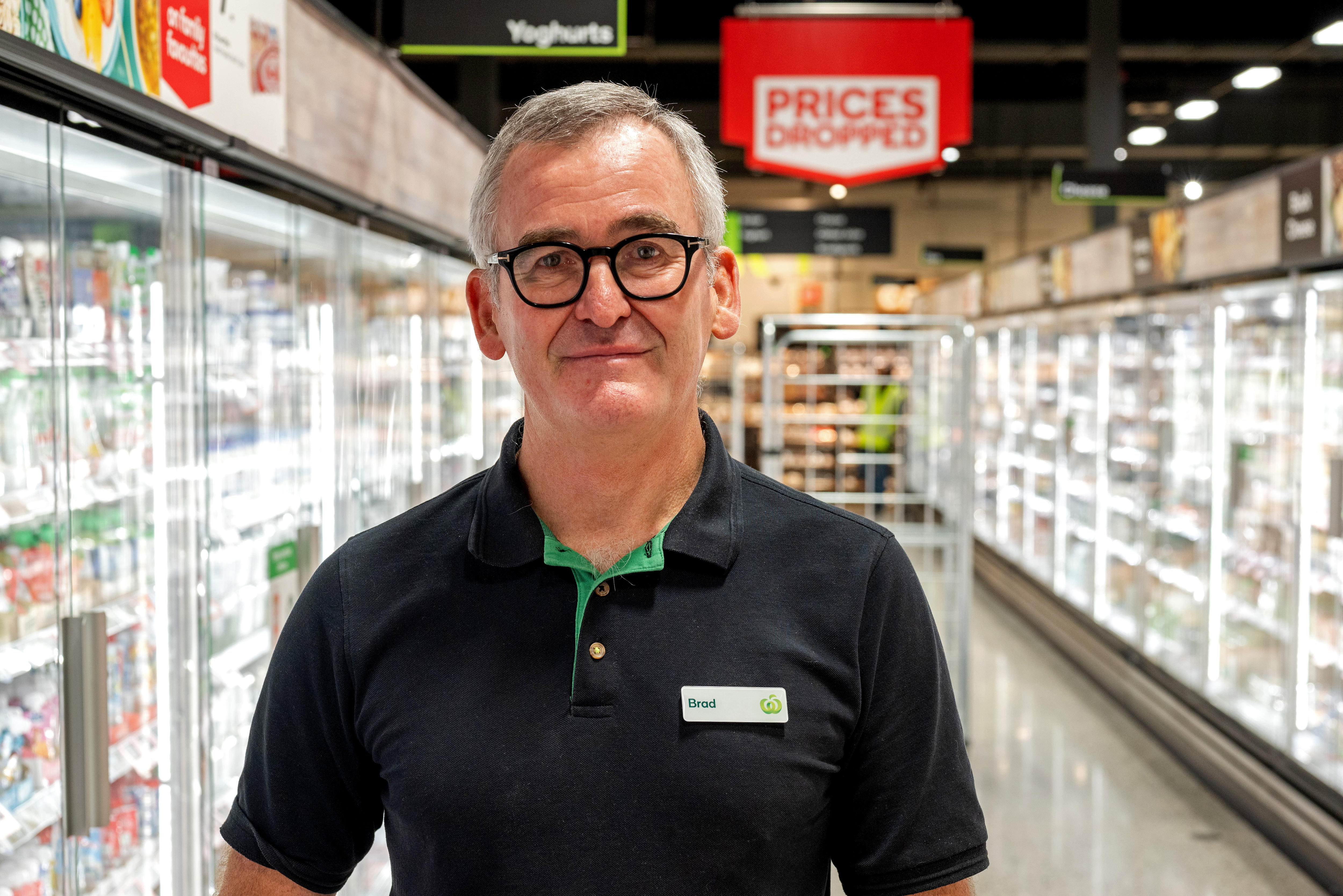 A man wearing a Woolworths polo shirt with the name tag 'Brad' stands in the cold food aisle of  supermarket.