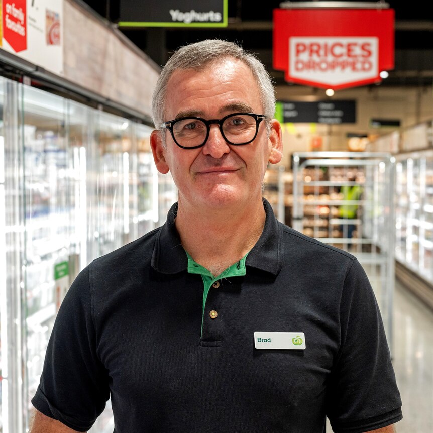 A man wearing a Woolworths polo shirt with the name tag 'Brad' stands in the cold food aisle of  supermarket.