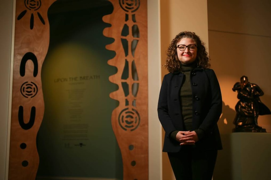 A woman in her 30s stands smiling happily in front of a doorway shaped with indigenous design.
