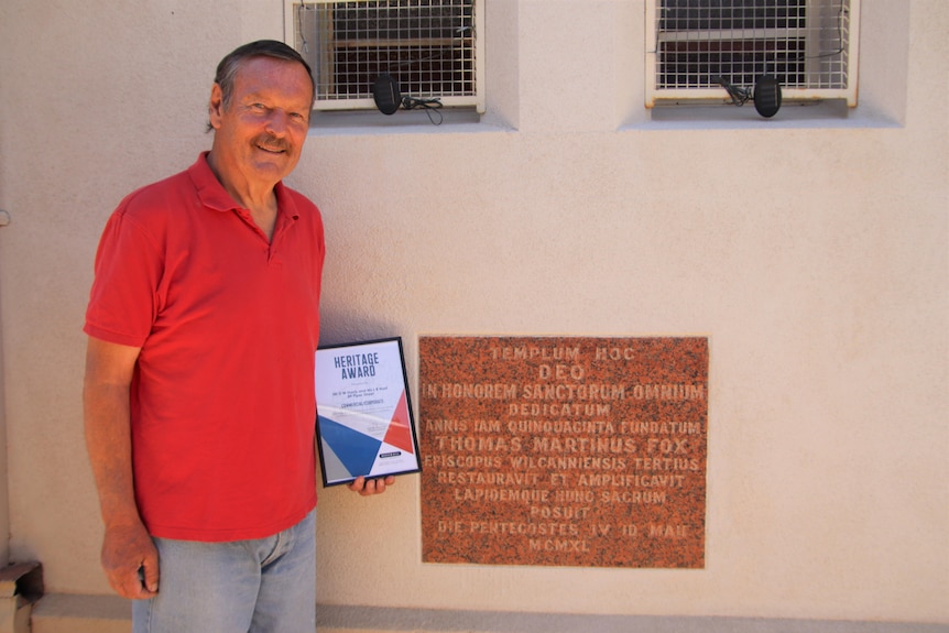 A smiling older man wearing a red shirt holding a plaque standing next to a wall with an inscribed foundation stone. 