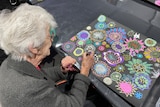 Elderly woman holds pen and draws bright colourful flowers on a large black piece of paper.