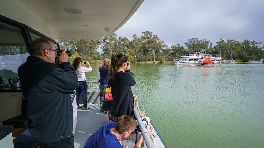 A group of young and old people take photographs from the deck of a paddle steamer on a river.