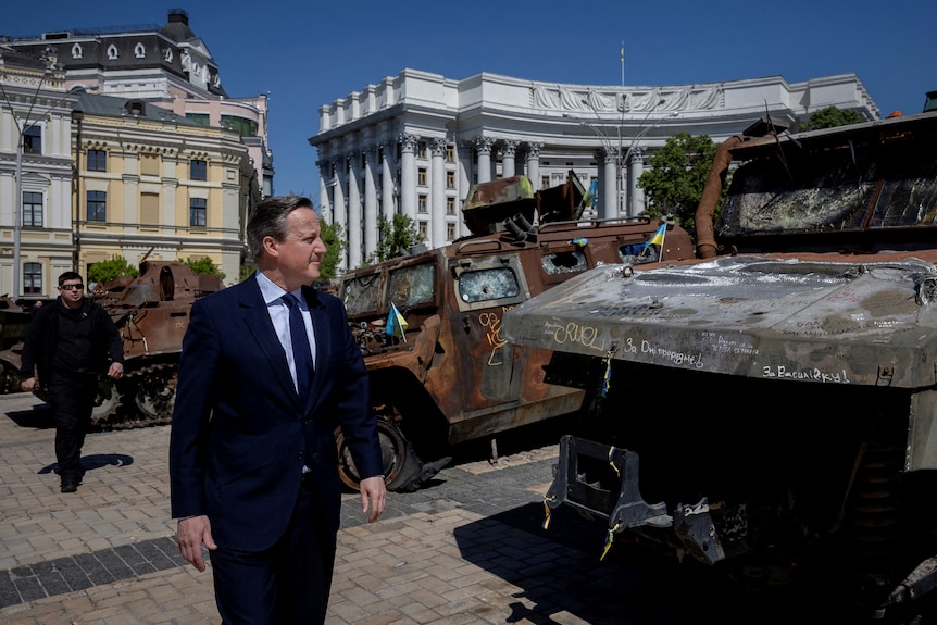 David Cameron wearing a suit walks past a destroyed van. 
